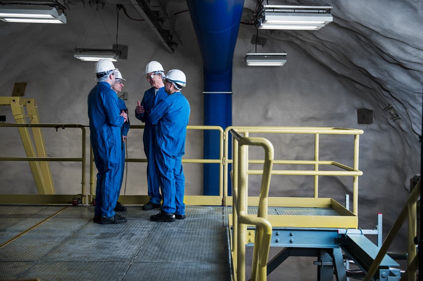 An image by photographer Luther Caverly of four men in blue coveralls with white hardhats and goggles conversing on scaffolding in an underground lab.